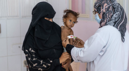 Une femme portant un voile noir lui couvrant le visage tient un enfant atteint de malnutrition sévère dans les bras, tandis qu'une infirmière mesure le diamètre du bras gauche de l'enfant.