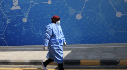 A nurse in full medical gear and a face mask walks on the street near a blue building with a cartoon image of a nurse. 