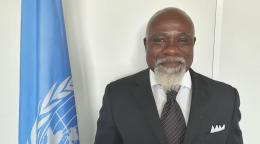 A man in a suit smiles with a UN flag to his side.