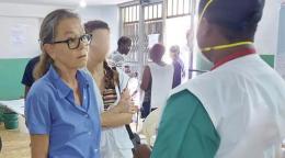 在海地的一家医院里，一名身穿蓝色上衣、戴着眼镜的妇女与一名从后面拍到的身穿白色长袍、戴着呼吸机面罩的护理人员交谈。这两名妇女被其他几个人包围着。