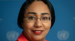 A woman in an orange coloured suit wearing glasses standing in front of a blue background with the UN logo