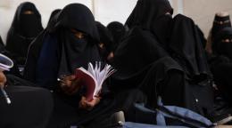 A row of women in full black clothing, sit on the floor with books laid out in front of them.