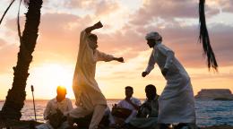 A group of men in white clothing dance together by a beachside as the sun sets in the background
