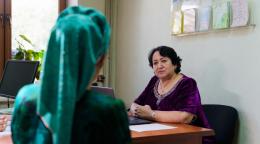 A woman in a green dress sits in a chair and talks to another woman in a purple dress in Tajikistan.