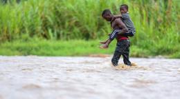 boy carries younger boy on his back as his walks through a river