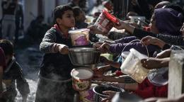 A young boy hands a large metal bowl, presumably with food, to people, including youth, lined up for food and water.