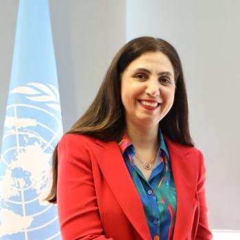 A woman with brown hair in a red blazer and blue shirt smiles at the camera with the light blue United Nations Flag behind her.