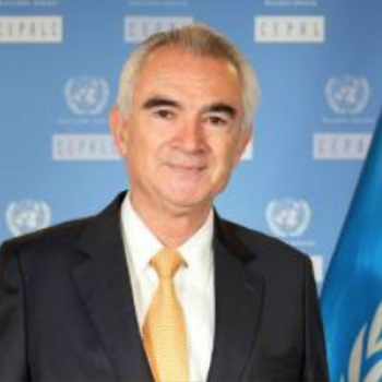 A man in a black suit and yellow tie stands in front of a blue background with UN logos.