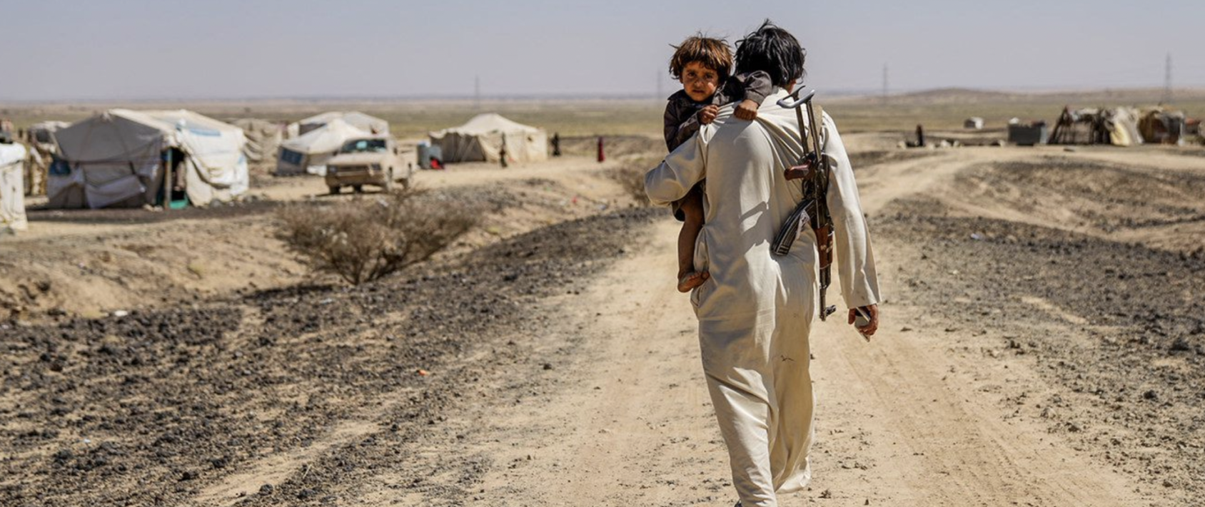 Un homme tient son enfant dans les bras et marche sur un chemin de terre, un fusil à l'épaule, vers un camp de réfugiés situé au milieu d'une étendue désertique.
