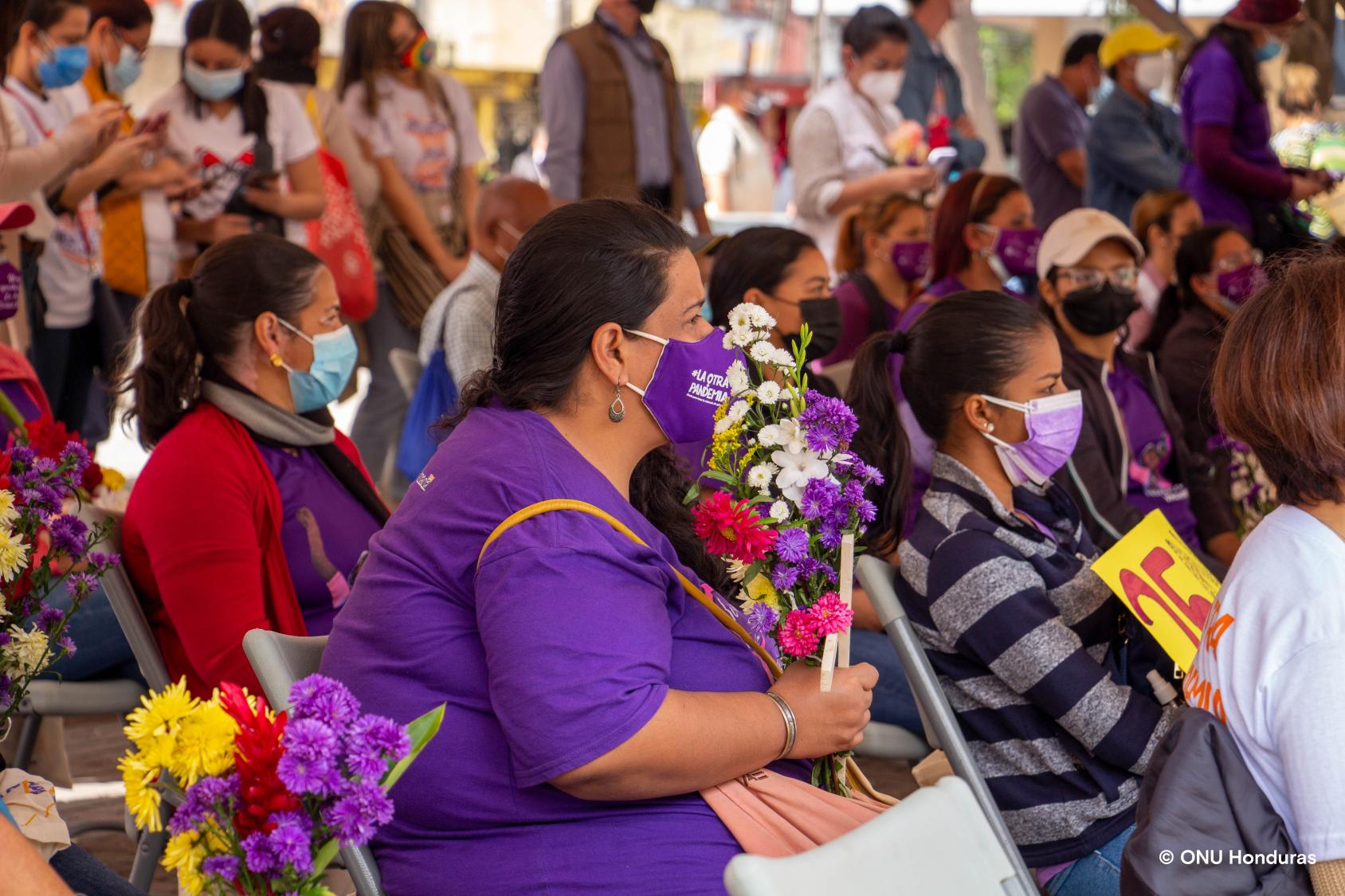 Las mujeres con mascarillas se sientan portando carteles, flores y cruces que representan a las víctimas de la violencia de género.
