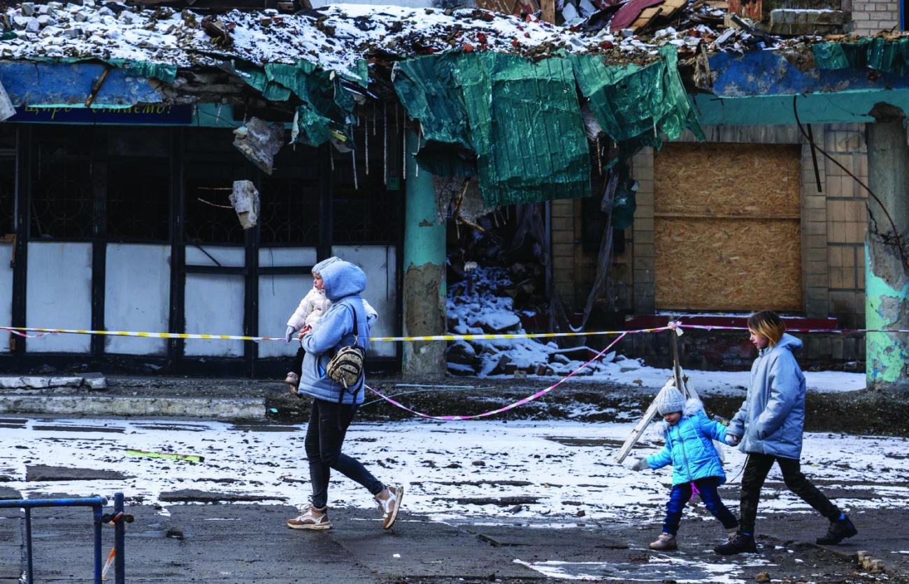 A woman walks in front of destroyed buildings and holds a child in her arms, with two children walking behind her.