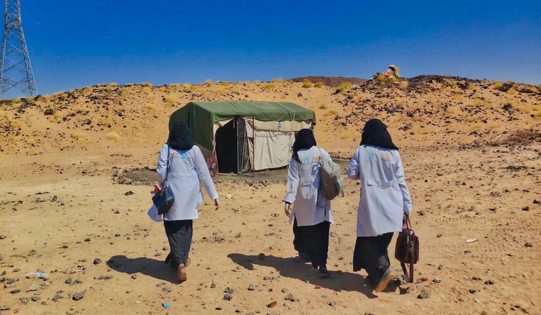 Three midwives wearing medical lab jackets walk away from the camera towards a mobile health clinic.