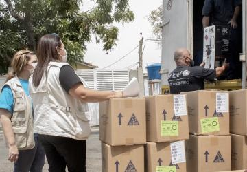 Un membre masculin du personnel de l’UNICEF portant un masque de protection aide à décharger des cartons dans le cadre de l’opération d’aide humanitaire réalisée au Venezuela. Deux femmes portant un masque de protection se tiennent debout près des cartons déchargés.