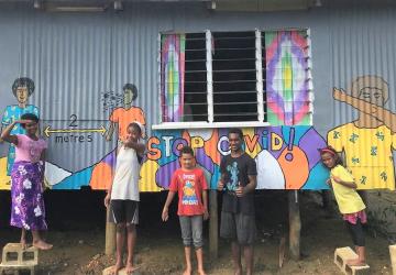 Des enfants se tiennent devant une peinture murale réalisée à la main. Ils sourient à la caméra avec fierté. 