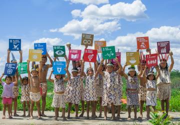 Young indigenous children wearing brightly coloured outfits proudly hold up SDG signage.