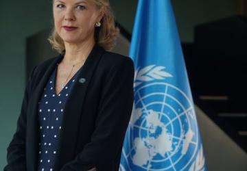 Una mujer con un vestido azul y una chaqueta negra se encuentra frente a la bandera de las Naciones Unidas.