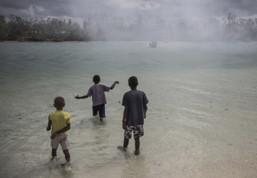 Trois enfants se tiennent debout dans un plan d'eau, dos à la caméra et regardent un bateau s’éloigner.