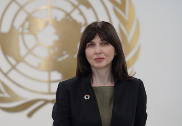 A woman, standing in front of the United Nations symbol, wearing a black jacket and green shirt looks directly at the camera. 