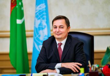  Мужчина в костюме улыбается в камеру, сидя на фоне флагов Туркменистана и Организации Объединенных Наций. 