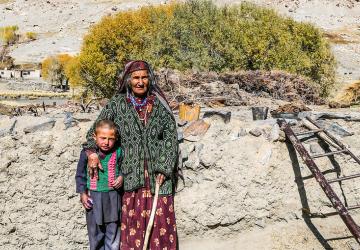 Une femme âgée se tient debout devant la caméra, le bras posé autour d'un jeune enfant. On aperçoit en arrière-plan un arbuste au milieu d'un paysage montagneux et désertique.