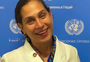 Portrait de Yesim Oruc, la nouvelle Coordonnatrice résidente des Nations Unies au Guyana, souriant à la caméra avec un panneau bleu et blanc des Nations Unies en arrière-plan.