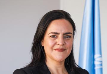 Una mujer con una chaqueta negra, de vestir, mira directamente a la cámara con la bandera de las Naciones Unidas detrás de ella.