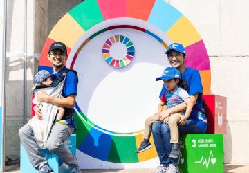 يحمل كل من الأب والأم طفلًا في أحضانهما بينما يبتسمان للكاميرا جالسين أمام عجلة أهداف التنمية المستدامة.
