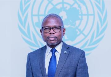 Retrato de un hombre con traje y corbata azul delante del emblema de las Naciones Unidas.
