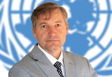 A man, in a grey suit, with his arms folded looks directly at the camera with a United Nations symbol in the background.