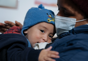 A baby in a blue hat rests on his mother who is wearing a face mask. 
