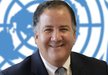 A smiling man looks straight at the camera with the United Nations Logo in the background.