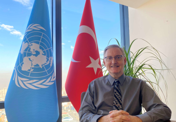 Foto oficial del recién nombrado Coordinador Residente para Turquía, Álvaro Rodríguez. Está sentado en un escritorio con las manos cruzadas y sonriendo delante de las banderas de la ONU y de Turquía.