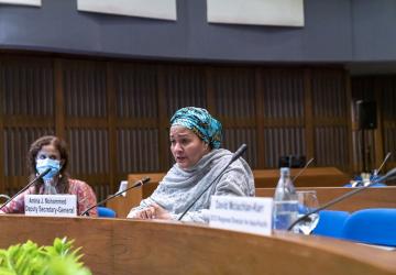 Une femme vêtue d'une robe de couleur claire et portant un turban s'exprime dans un microphone lors d'une réunion.