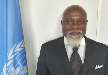 A man in a suit smiles with a UN flag to his side.