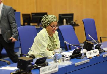 Une femme portant un châle jaune, un turban et des lunettes est assise à une table rectangulaire bleue, dans une salle de conférence, et parle dans un microphone lors d'une réunion organisée par l'ONU.