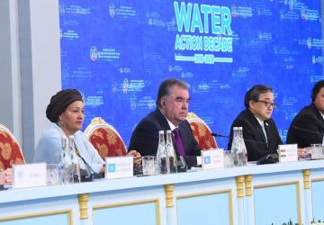 A woman sits on a plenary, with several men to both sides, also seated at microphones with a sign that says 'Water Action Decade' behind them.