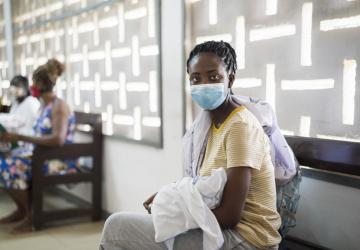 Une femme portant un masque de protection respiratoire et tenant son bébé dans les bras est assise dans une salle d'attente médicale avec d'autres femmes, le regard tourné vers l'objectif.