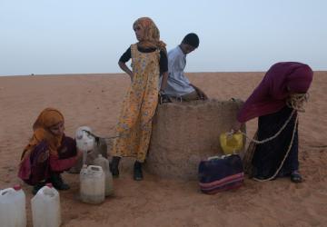 In a red-sanded desert, four young people try to fill up water jugs from a well.