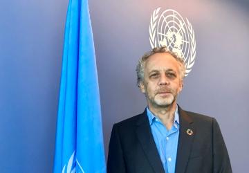 Un Coordinador Residente-funcionario de las Naciones Unidas se encuentra frente a una pared azul y una bandera azul de las Naciones Unidas.