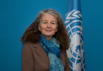 Une femme aux cheveux longs, bouclés et grisonnants et portant un blazer marron et une écharpe bleue se tient près d'un drapeau de l'ONU et regarde la caméra en souriant.