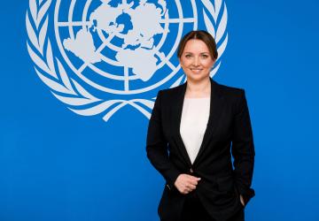 Woman in a black suit stands against a blue background with the UN logo