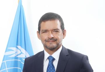 Man in a dark blue suit and blue tie standing in front of a light blue UN flag