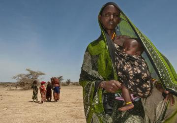 A woman holds a child in her arms, amid arid, dusty land that looks parched and dry.