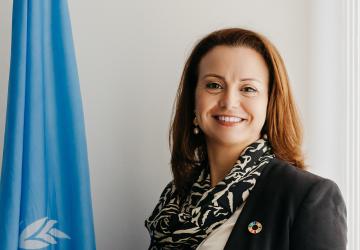 A woman with brown hair and a black suit and scarf, the UN Resident Coordinator in UAE, stands next to a blue UN flag.