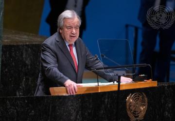 A man, the UN Chief, Antonio Guterres, in a black suit and red tie, stands at a podium against a blue background speaking into a microphone