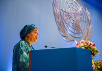 A woman in a green dress and headscarf speaks on a blue podium