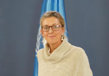 A woman in a white sweater and glasses standing in front of a blue flag with the UN logo on it.