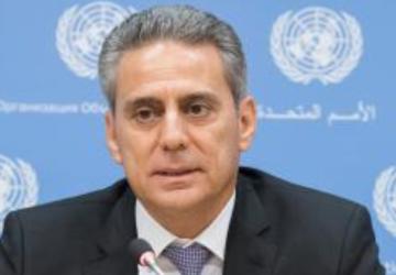 Muhannad Hadi sits in front of a lectern wearing a black suite and grey tie and behind him is a screen with UN logos