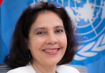 A woman in a white blazer sitting in front of a blue UN flag