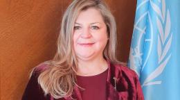 Official photo shows Catherine smiling to the camera, wearing a wine coloured jacket and blouse. She stands by the UN flag.